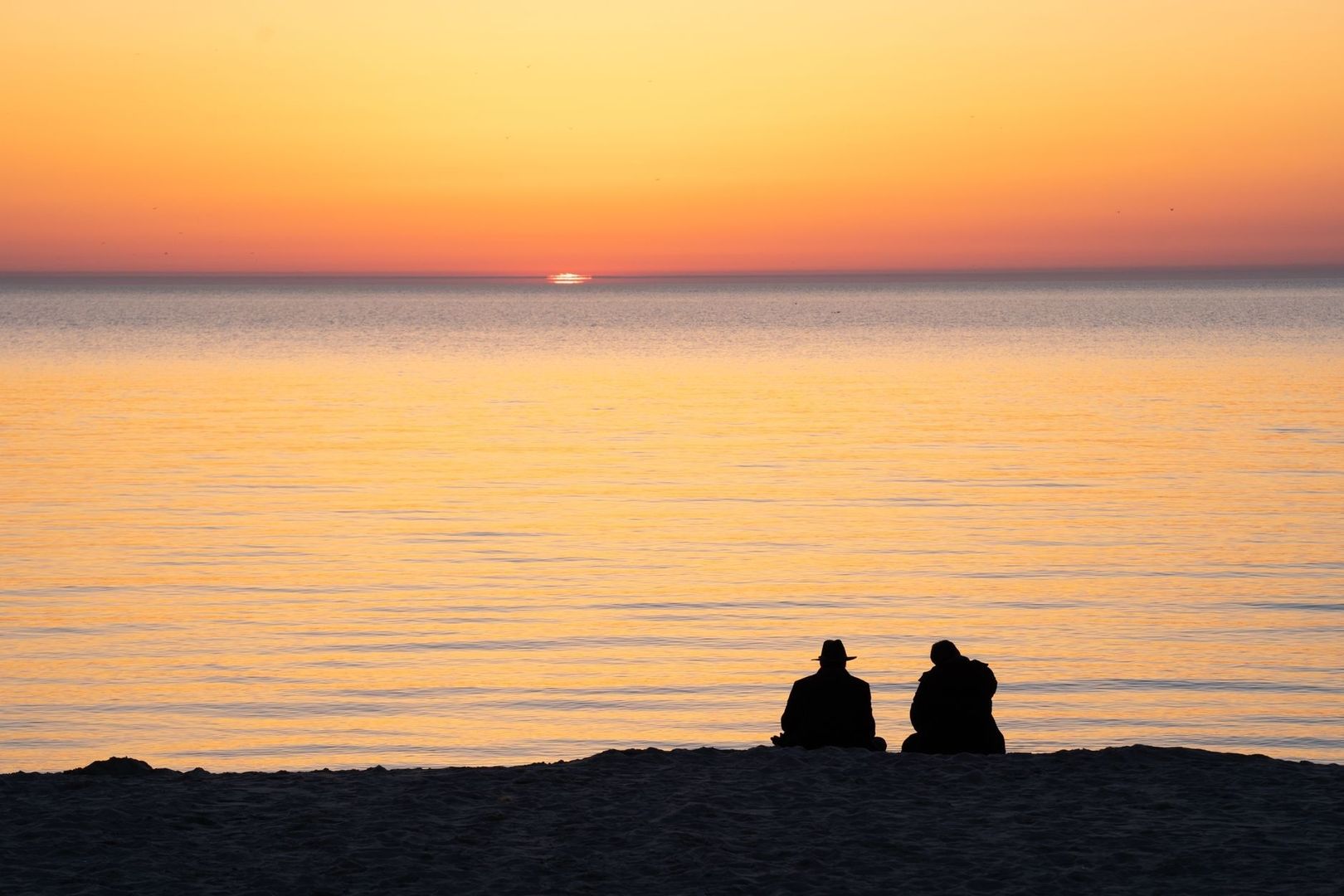 Kaksi ihmistä istuu rannalla katsomassa auringonlaskua